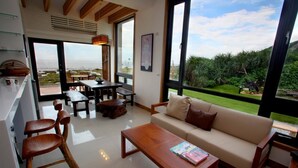 Una sala de estar moderna con un sofá, una mesa de centro y sillas de madera. Amplias ventanas ofrecen vistas al exterior.