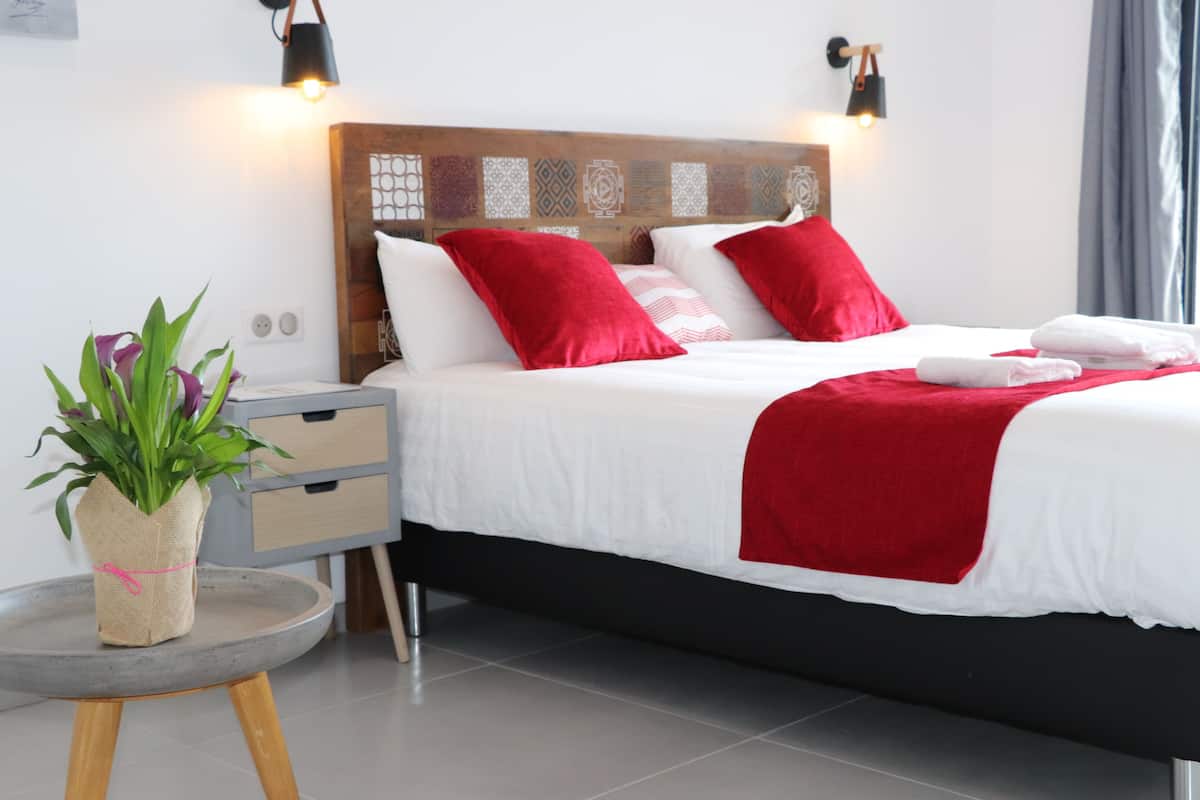 A neatly made bed with red and white bedding, a small round table with a plant, and a nightstand with drawers.
