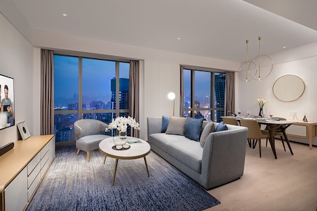 A modern living room with a sofa, coffee table, and dining area, featuring large windows with a city view.