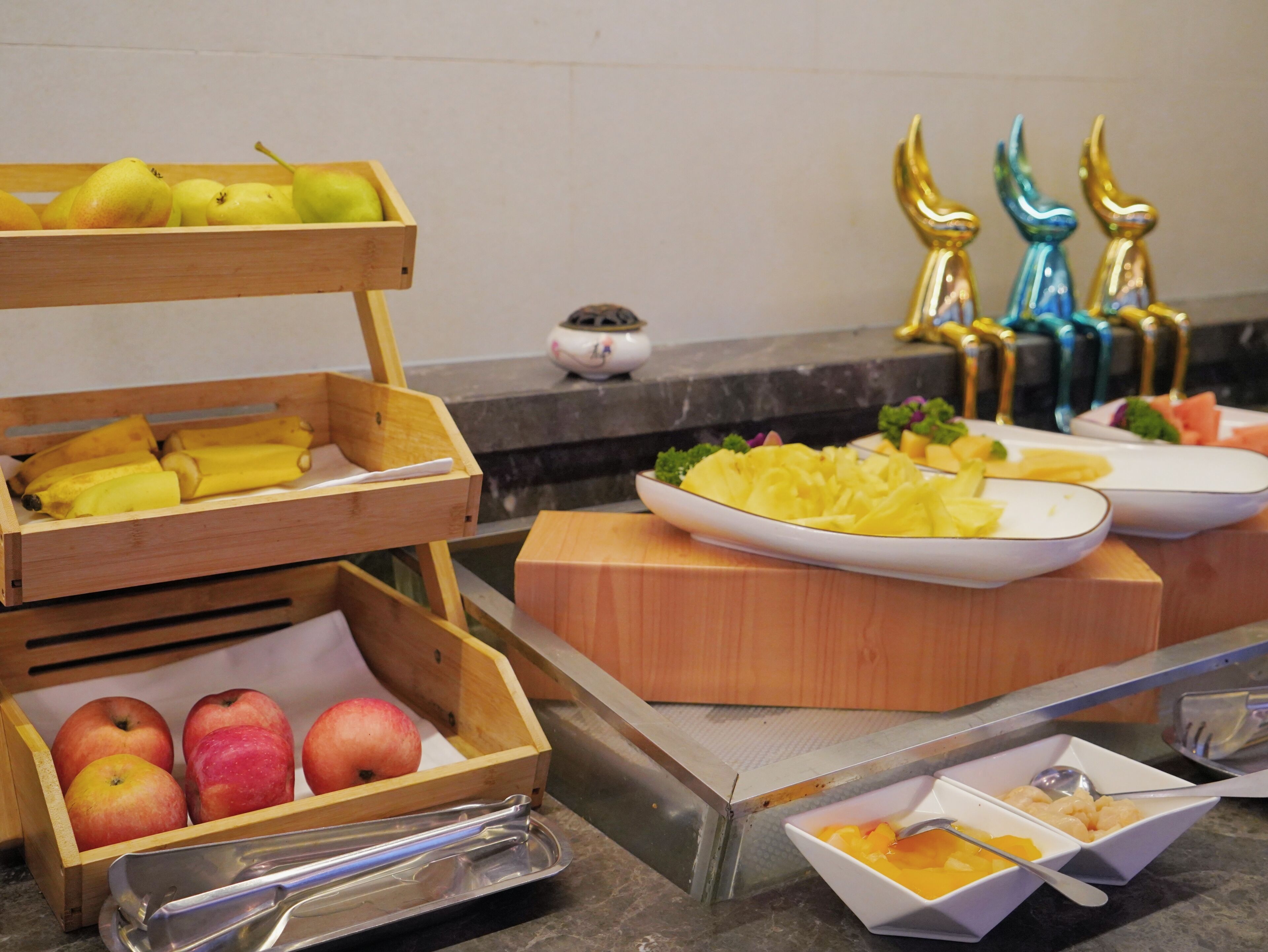 A buffet with wooden fruit and vegetable trays, a white plate with pineapple chunks, and a white bowl with a spoon.