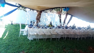 Un montaje para comedor al aire libre con una larga mesa cubierta con un mantel blanco, rodeada de sillas de plástico transparente, bajo una pérgola.