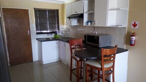 A compact kitchen with a microwave, two wooden chairs, a small dining table, and a window with blinds. - Ranks Guesthouse (Molepolole)