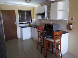 Cocina compacta con microondas, dos sillas de madera, una mesa pequeña y una ventana con persianas.