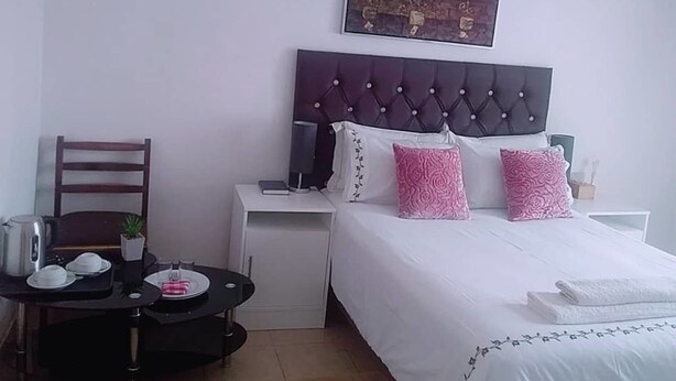 A bedroom with a bed, a nightstand, a bedside table, a chair, and a small table with a plant and a teapot. - Ranks Guesthouse (Molepolole)