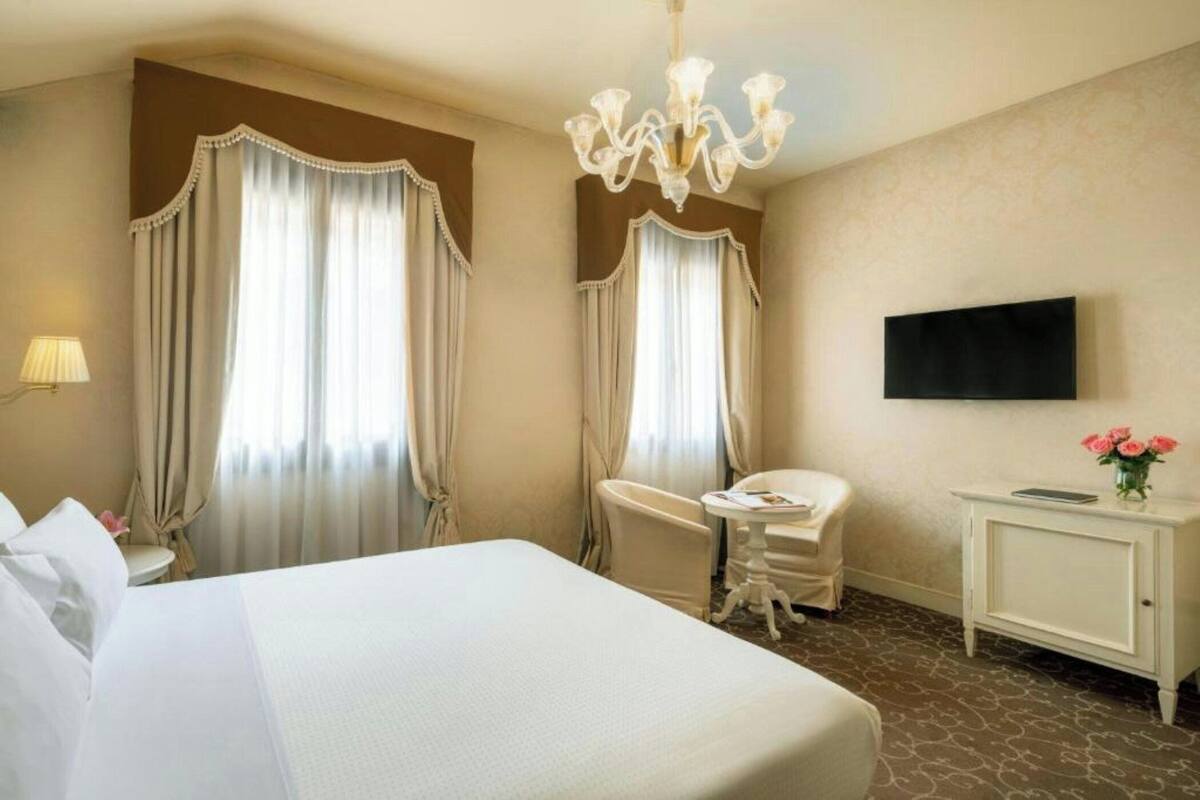 A hotel room with a large bed, a desk, a chair, a television, and a chandelier.