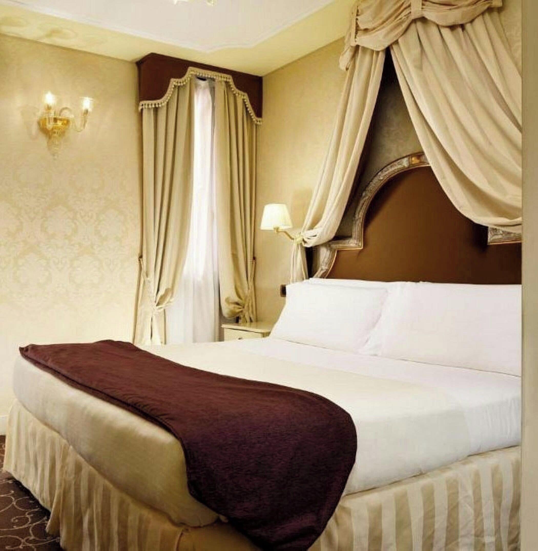 A hotel room with a large bed, curtains, a wall-mounted lamp, and a patterned wall.