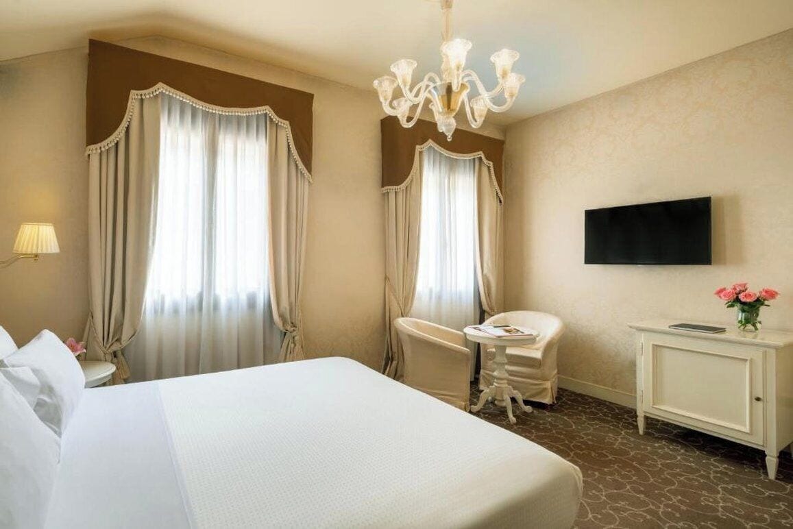 A hotel room with a large bed, a desk, a chair, a television, and a chandelier.