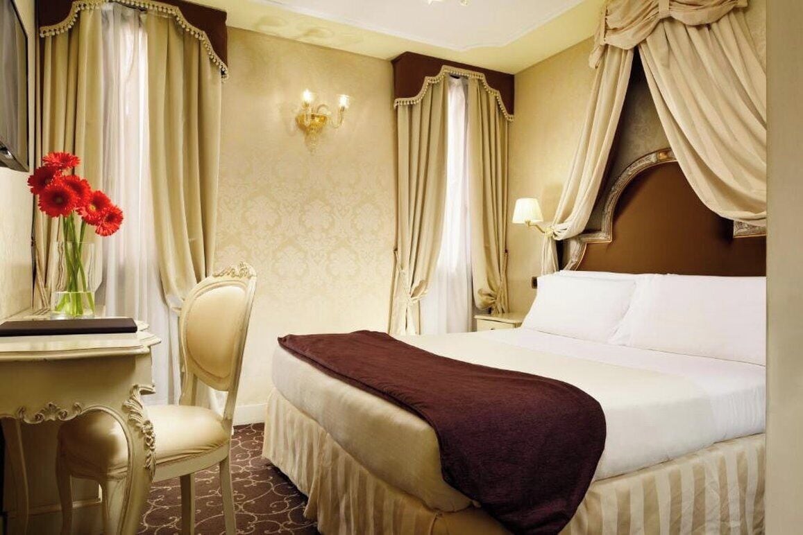 A hotel room with a bed, a desk, a chair, a vase with flowers, and curtains.