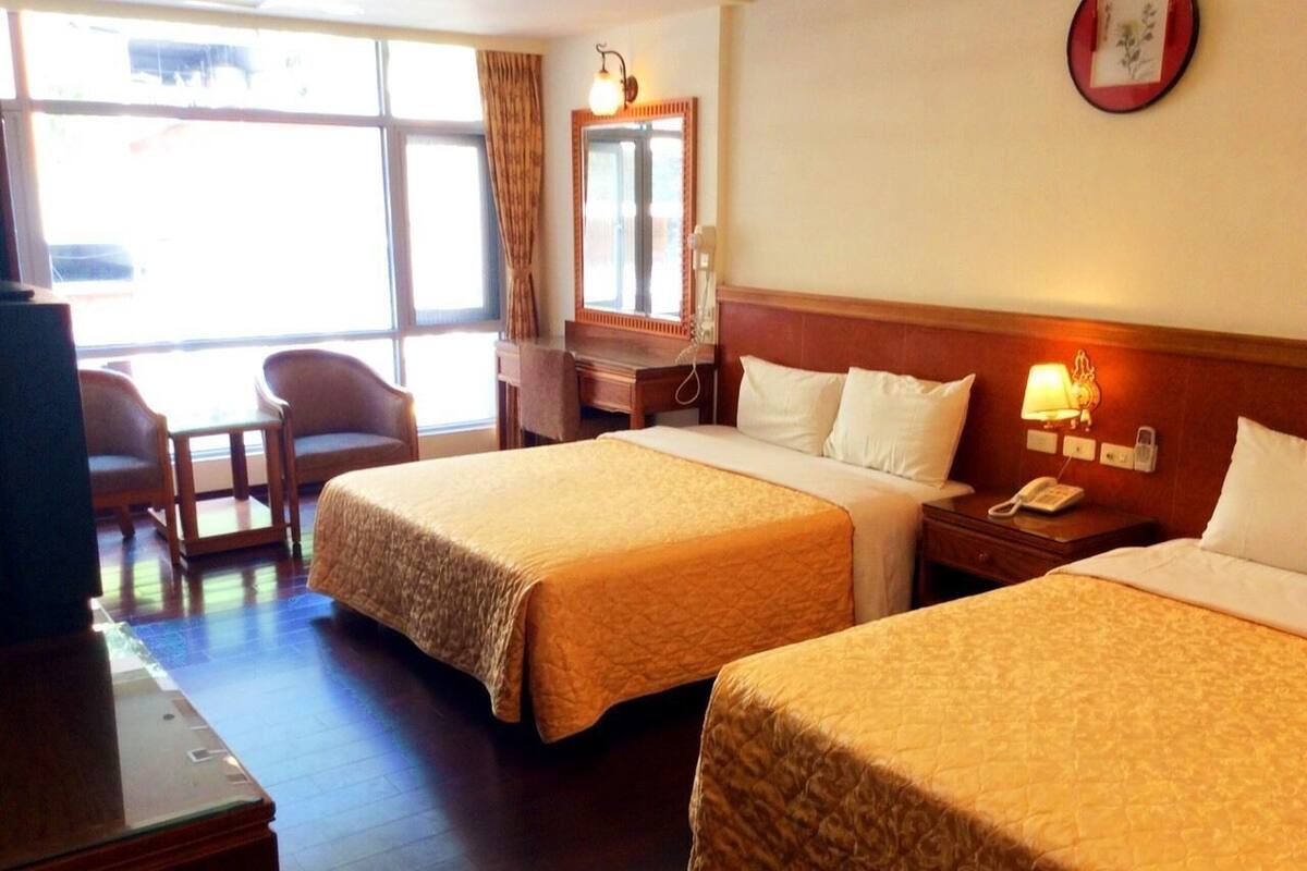 A hotel room with two beds, a desk, a chair, a mirror, and a window.
