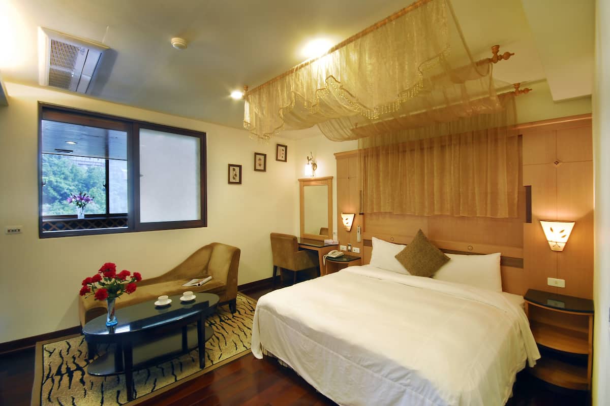A hotel room with a large bed, a desk with a chair, a small table with a vase of flowers, a mirror, and a window with a view of trees.