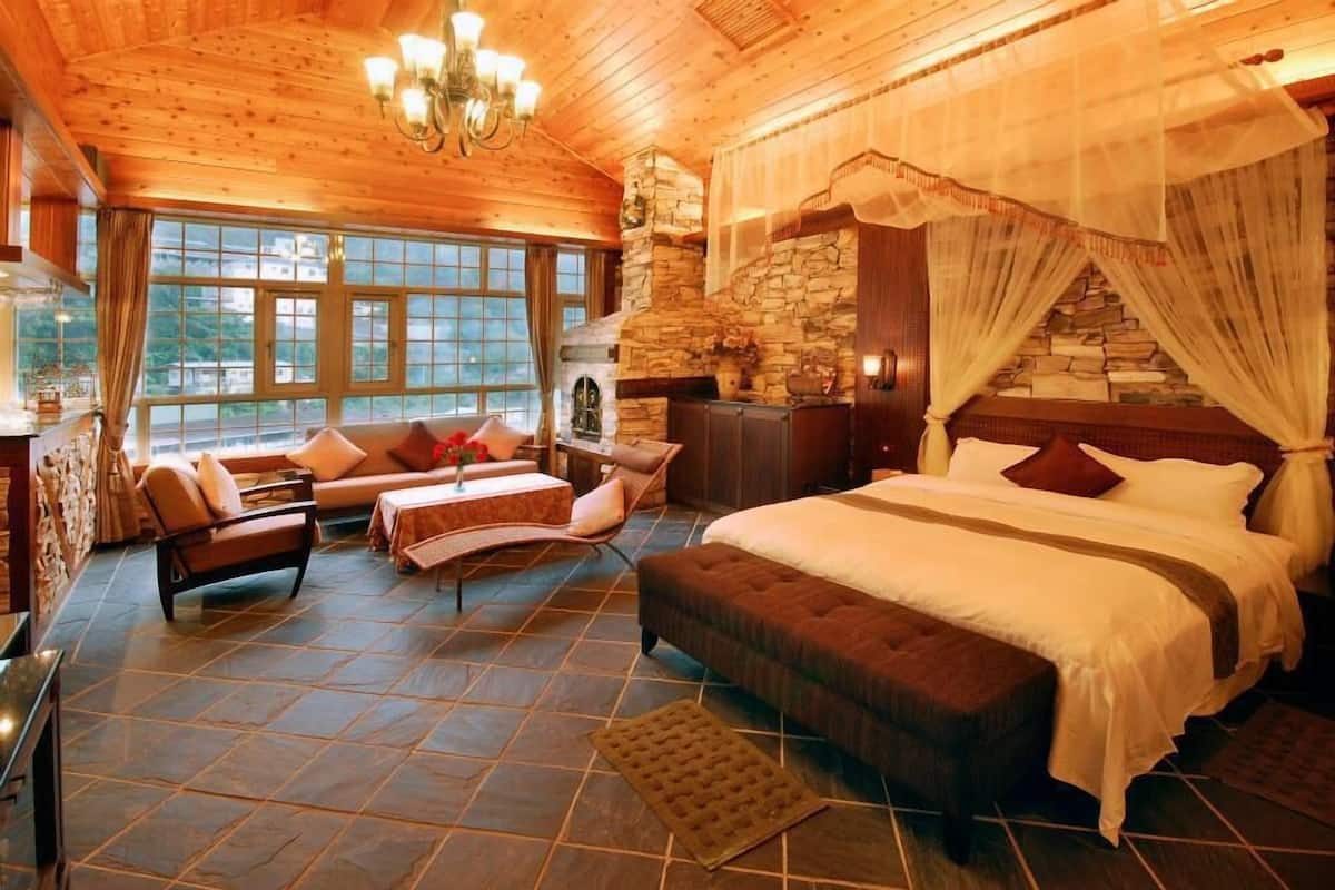A spacious bedroom with a large bed, a stone fireplace, and a view of the outdoors through large windows.