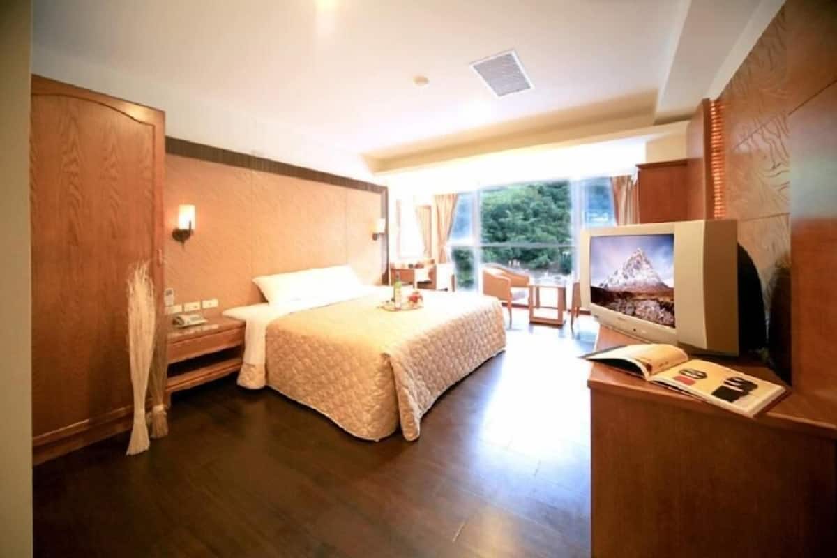 A hotel room with a large bed, a desk with a computer, a television, and a window with a view of trees.