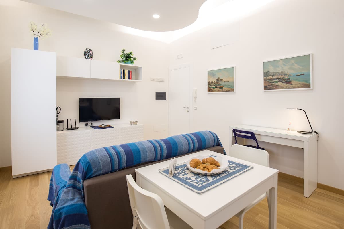 A modern living room with a sofa, a dining table with a plate of croissants, a TV, and a desk with a lamp.