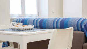 Un'area pranzo con un tavolo e sedie bianche, un cuscino a righe blu e grigio e una finestra con persiane.