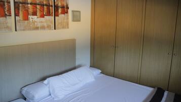 A neatly made bed with white linens, a wooden wardrobe with two doors, and a framed abstract painting on the wall.