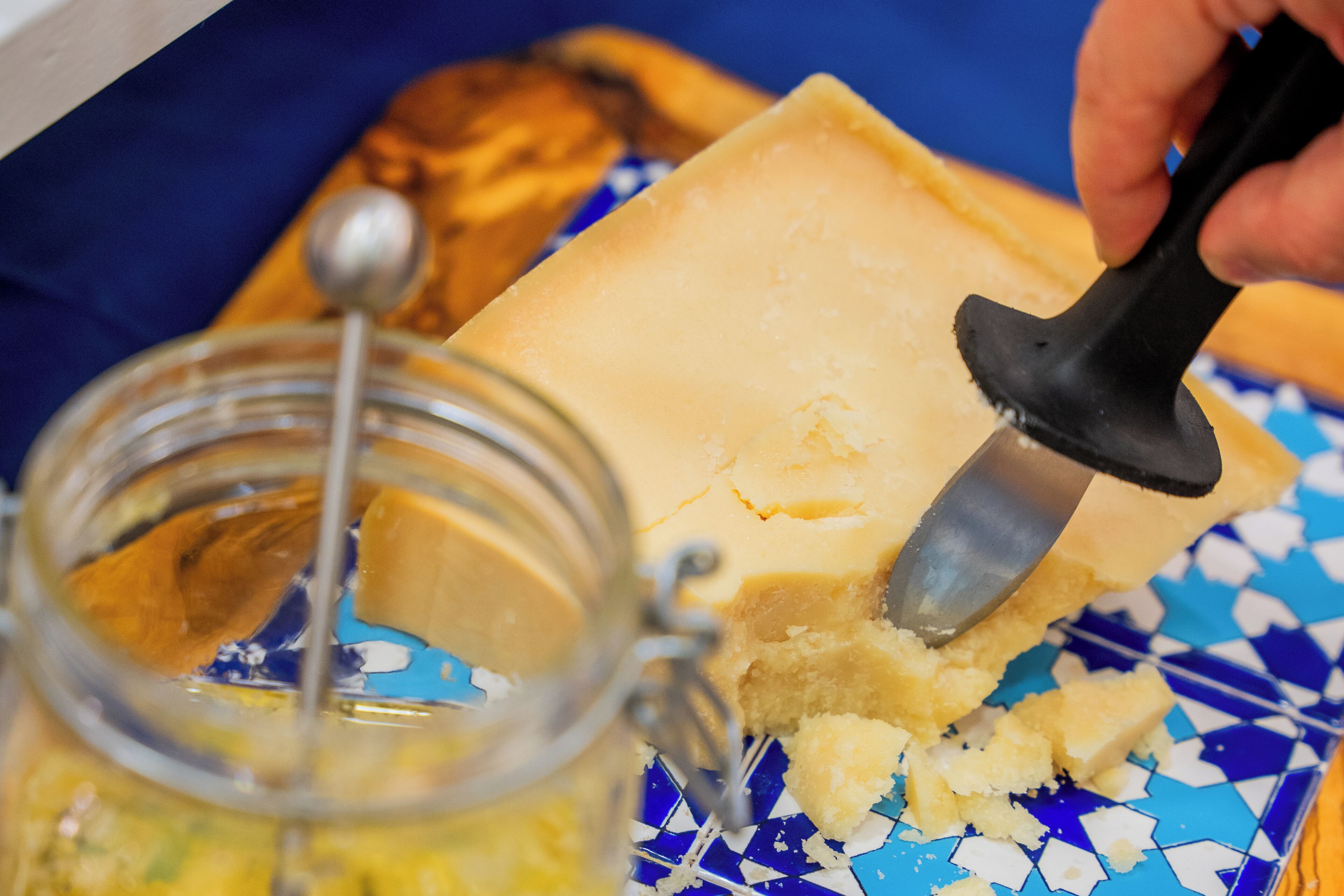Close-up de uma mão usando um fatiador de queijo em um pedaço de queijo, com um pote de molho por perto.