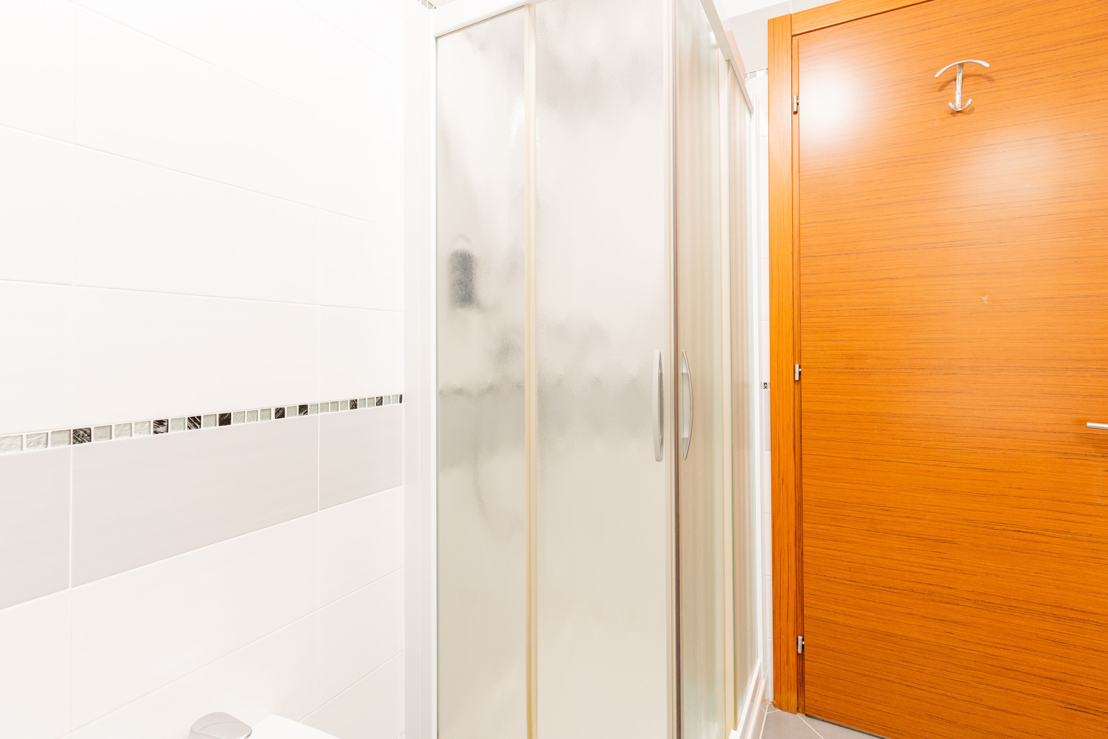 A bathroom with a glass shower enclosure, a wooden door, and white tiled walls.