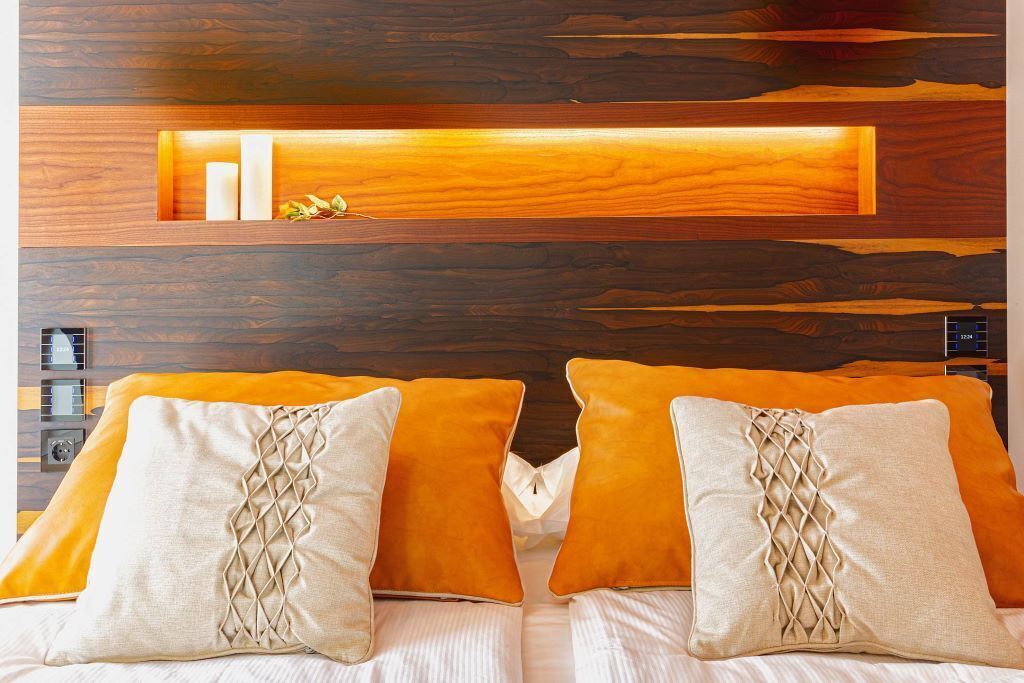 Een bed met twee oranje kussens en een beige kussen met een kruispatroon, voor een houten hoofdbord met een plank erboven.