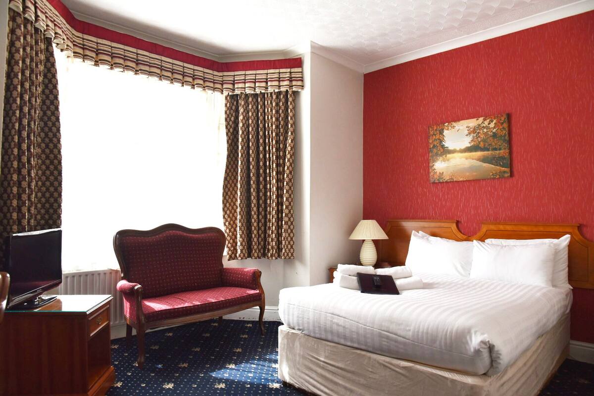 A hotel room with a bed, a desk with a computer, a red armchair, and a painting on the wall.