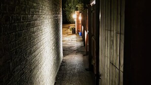 A narrow alleyway with brick walls and wooden doors, leading to a brighter area with a tree and a blue bucket. - Crystal Hotel (Cambridge)