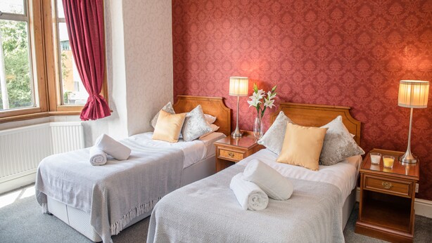 A hotel room with two beds, a window with red curtains, a radiator, and a patterned wallpaper. - Crystal Hotel (Cambridge)