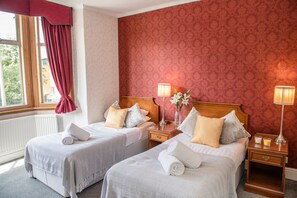 A hotel room with two beds, a window with red curtains, a radiator, and a patterned wallpaper. - Crystal Hotel (Cambridge)