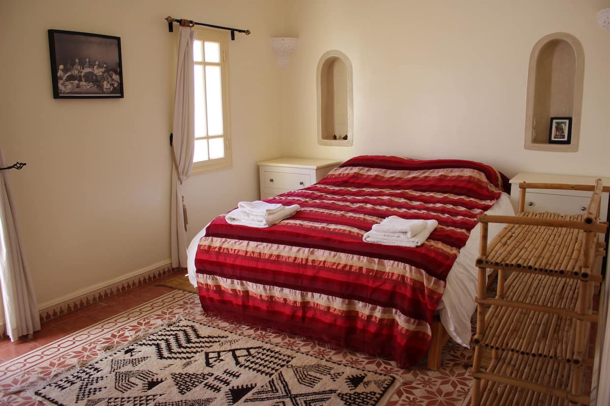 Un dormitorio con cama, mesita de noche, espejo y ventana con cortinas.