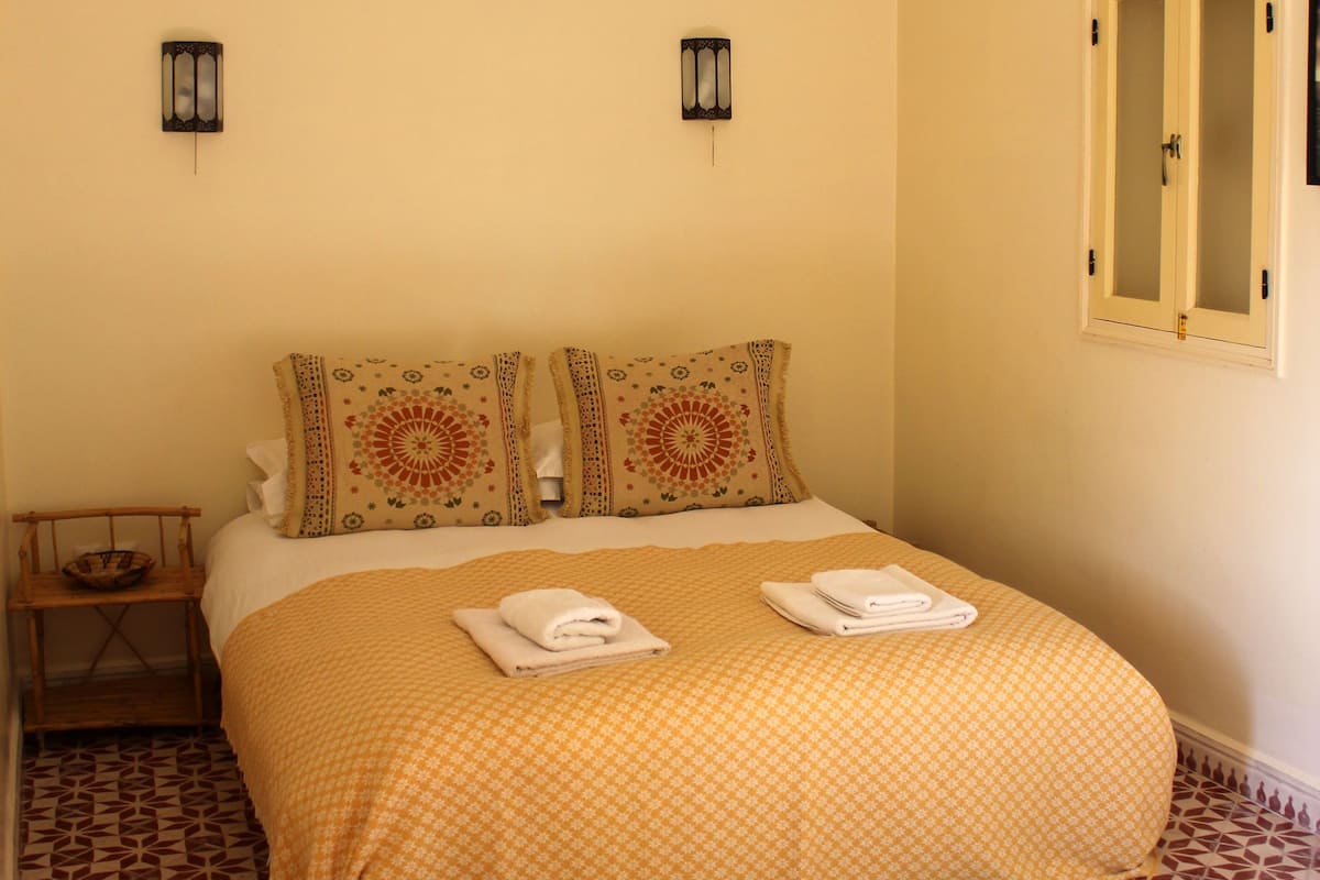 Una cama bien hecha con una colcha estampada, dos almohadones decorativos y dos toallas dobladas sobre la cama.
