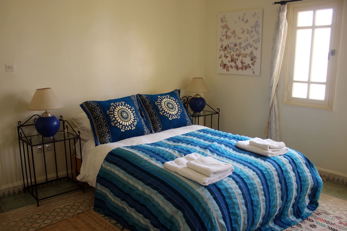 Una cama bien hecha con una colcha azul y blanca a rayas, dos almohadas iguales y dos lámparas de noche sobre pedestales de metal negro.