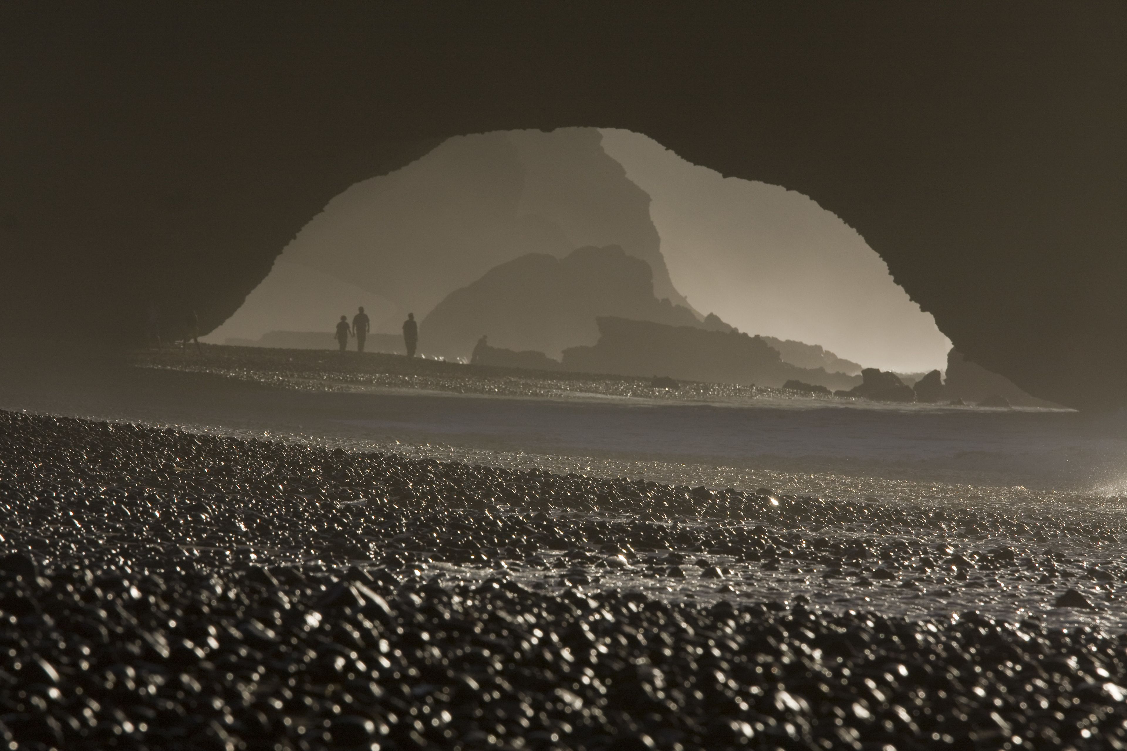 A beach with a large rock formation in the background and people walking on the shore.