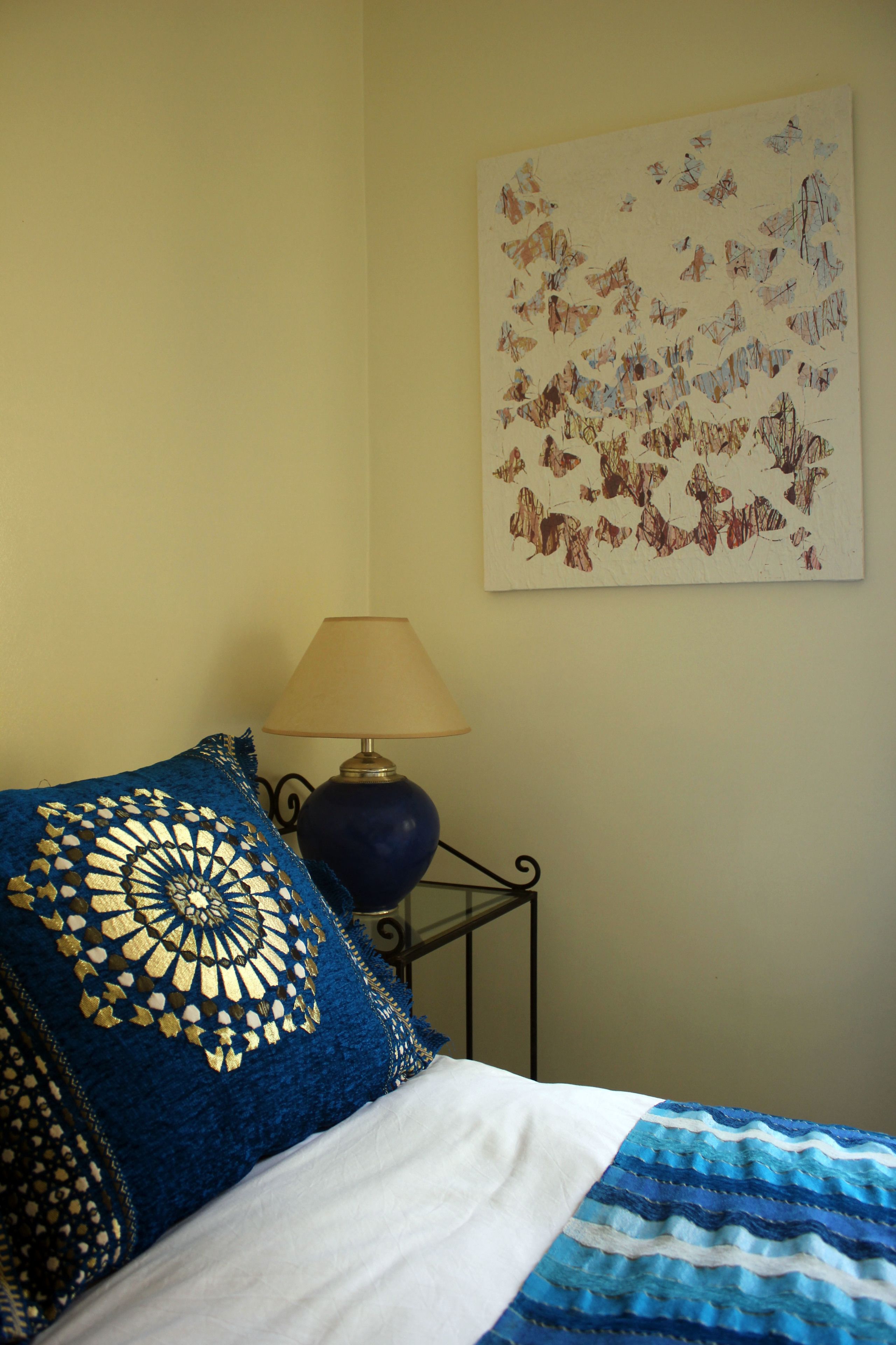 A bed with a blue and white blanket, a blue crocheted pillow, a lamp, and a wall with a butterfly painting.