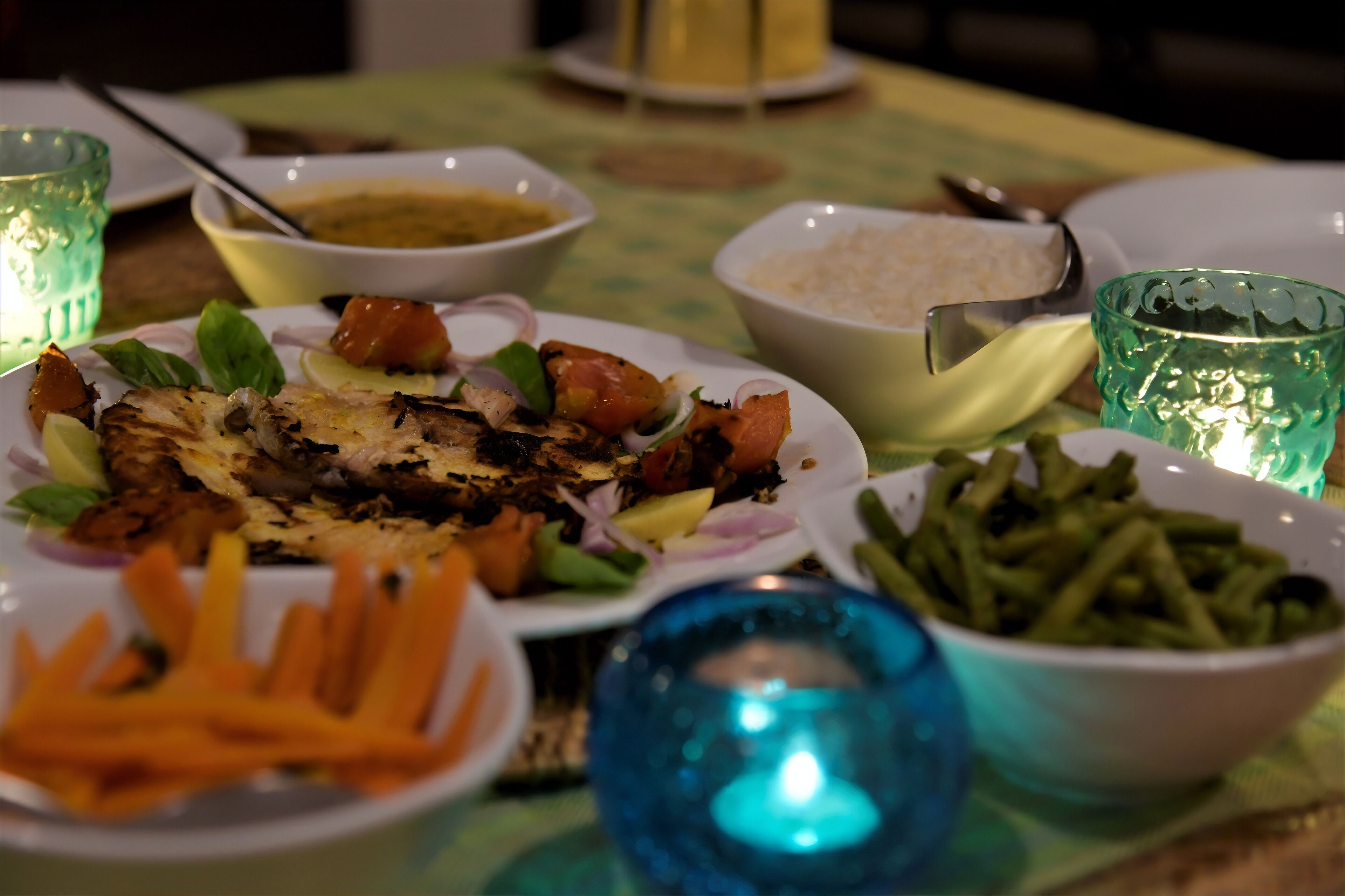 Una mesa dispuesta con diversos platos, incluyendo carne a la parrilla, verduras y arroz, con velas y cristalería.