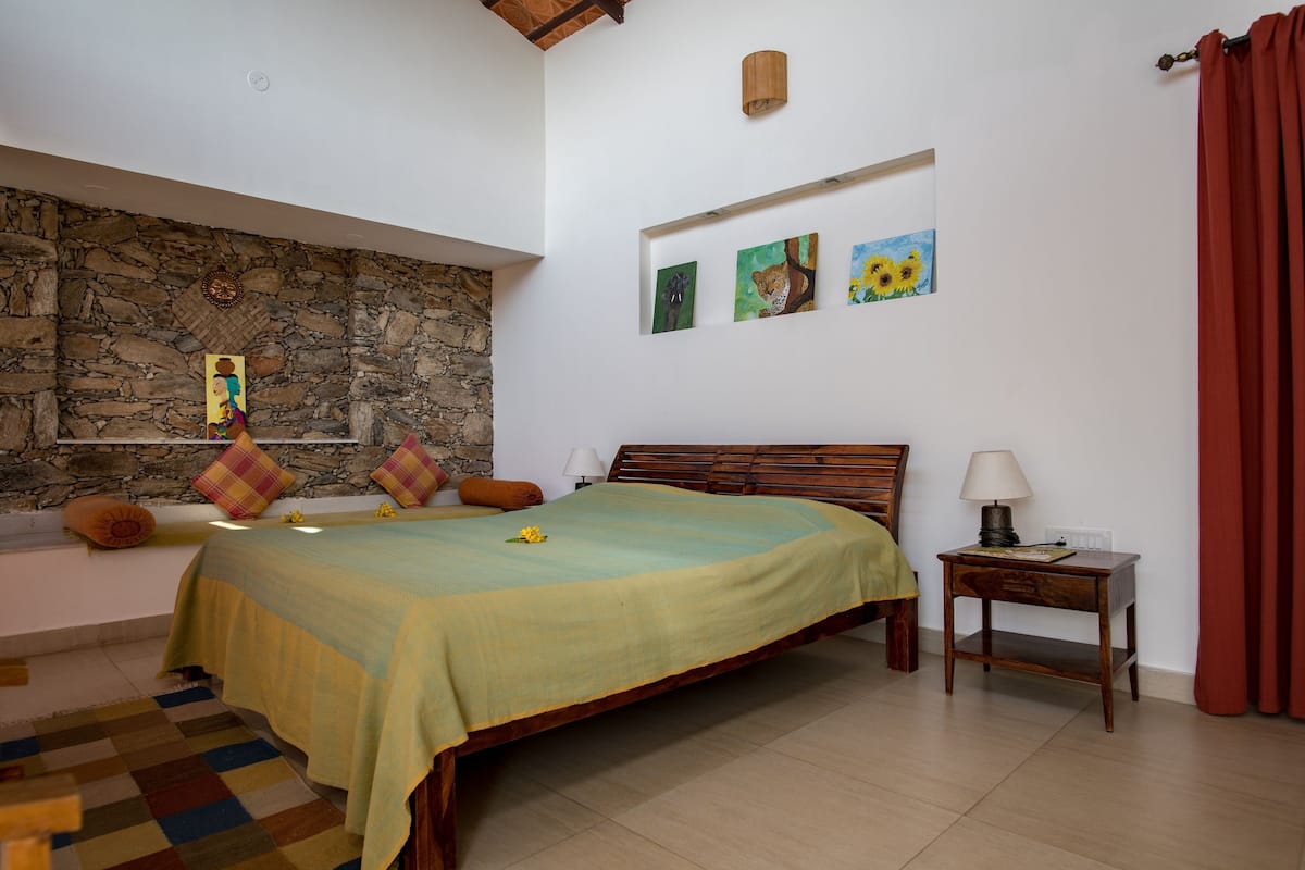Une chambre avec un mur en pierre, un lit recouvert d’une courtepointe verte, une tête de lit en bois, une table de chevet avec une lampe et une fenêtre ornée de rideaux rouges.