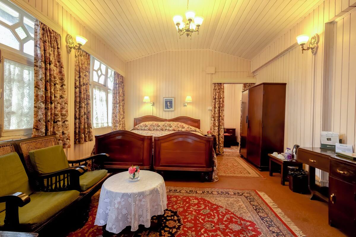 Une chambre traditionnelle avec un lit en bois, un canapé vert, une table ronde ornée d’un vase et un tapis rouge à motifs.