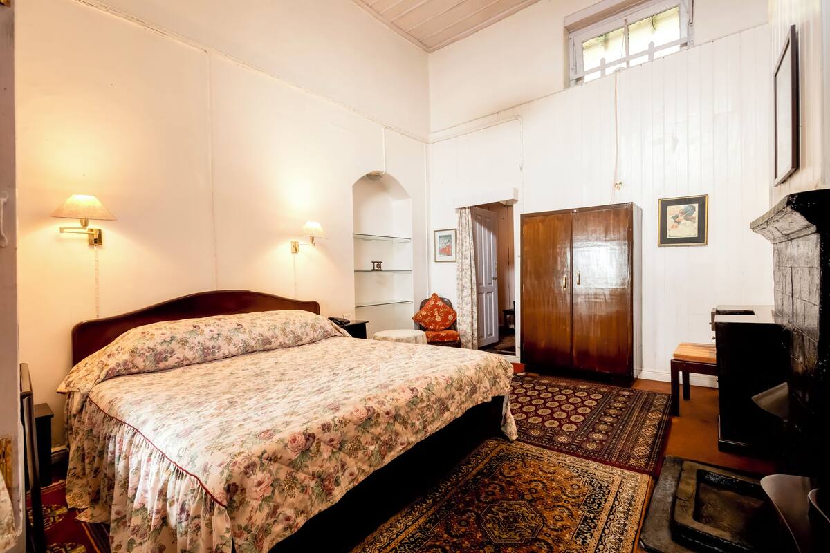 Une chambre avec un grand lit, deux lampes fixées au mur, une armoire en bois, une petite table et un tapis à motifs.
