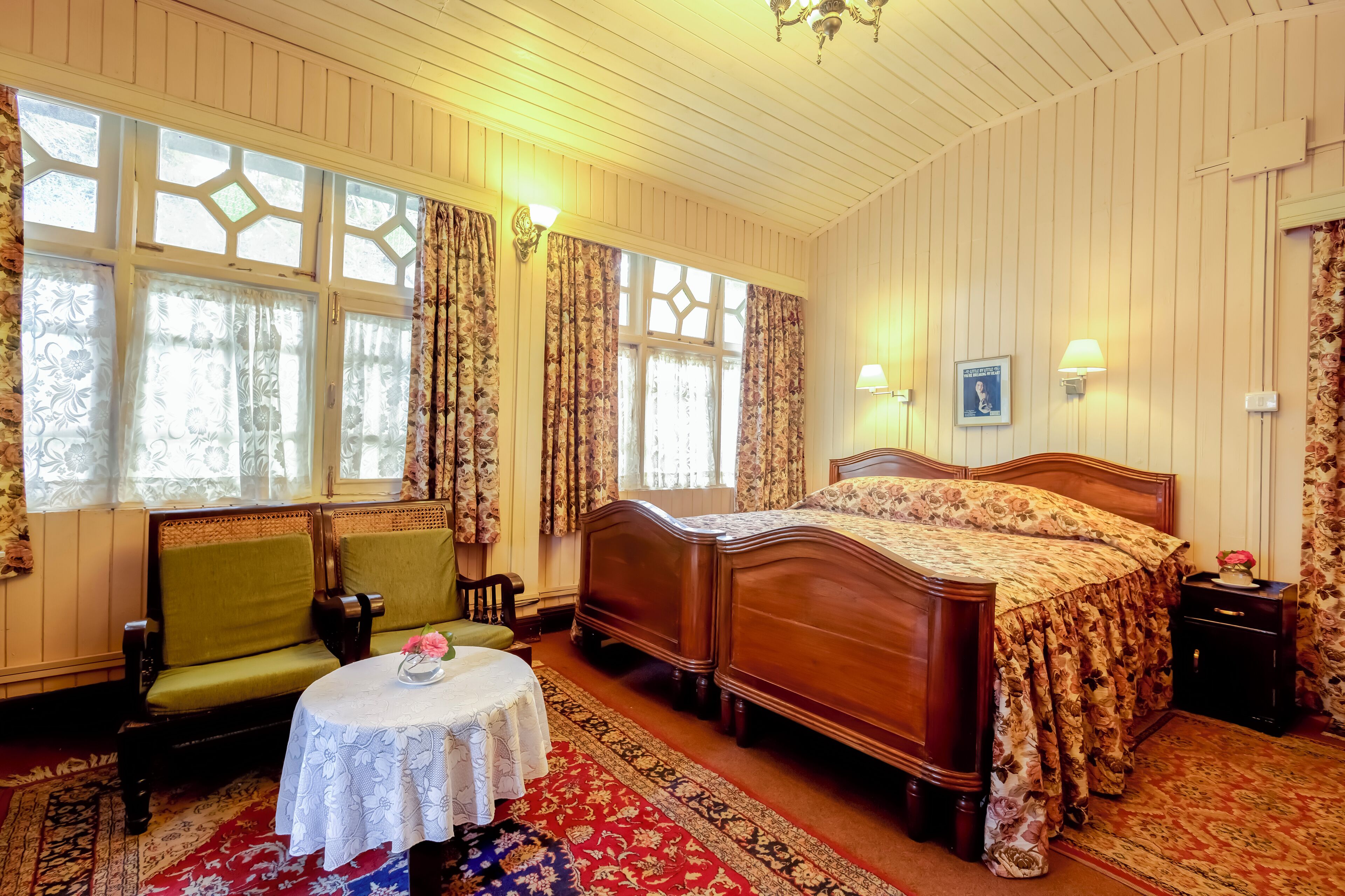 Een klassieke slaapkamer met een houten bed, een groene fauteuil, een klein rond tafeltje met een bloem en een kamerkleed met een patroon.