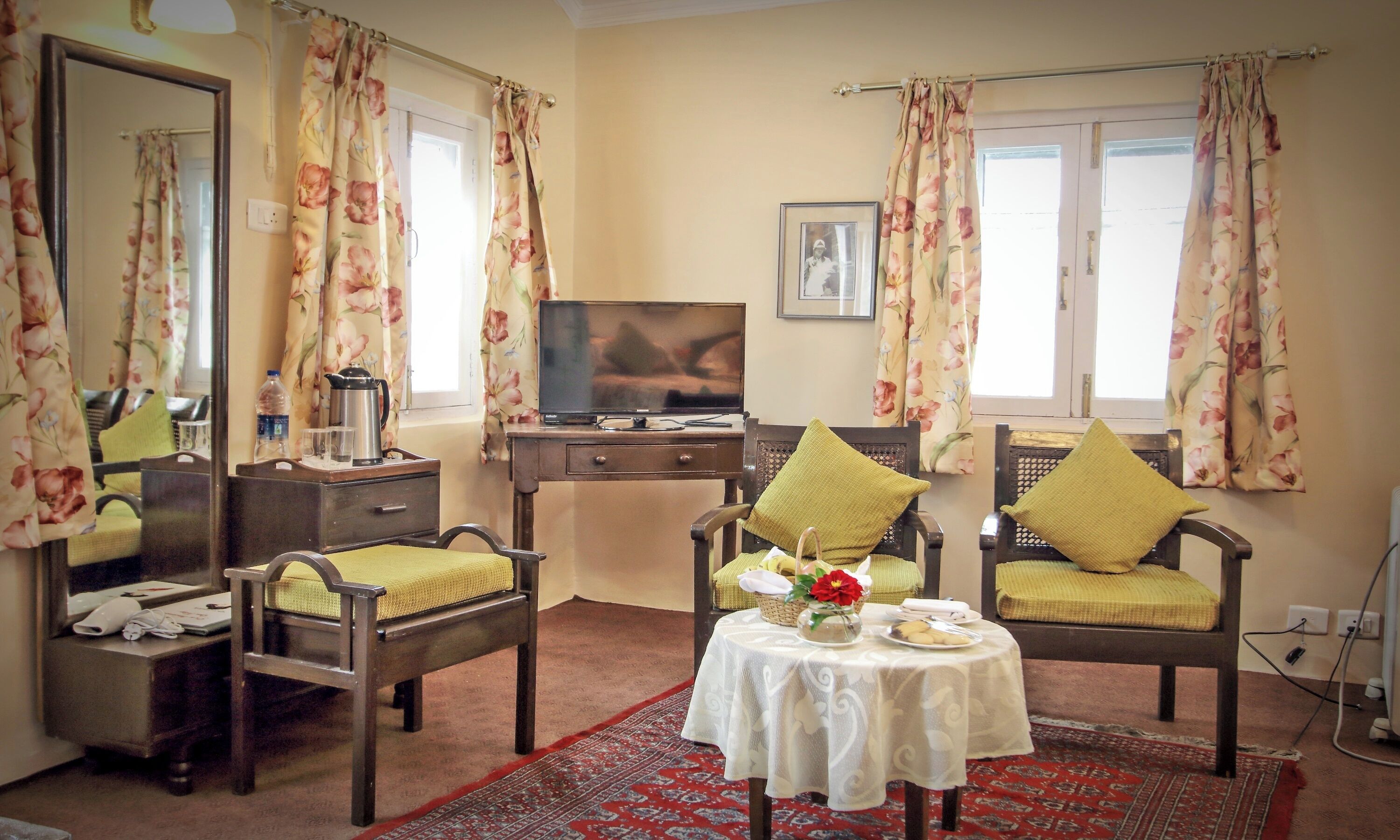 A hotel room with a television, two wooden chairs with cushions, a small table with a vase and a plate, and floral curtains.