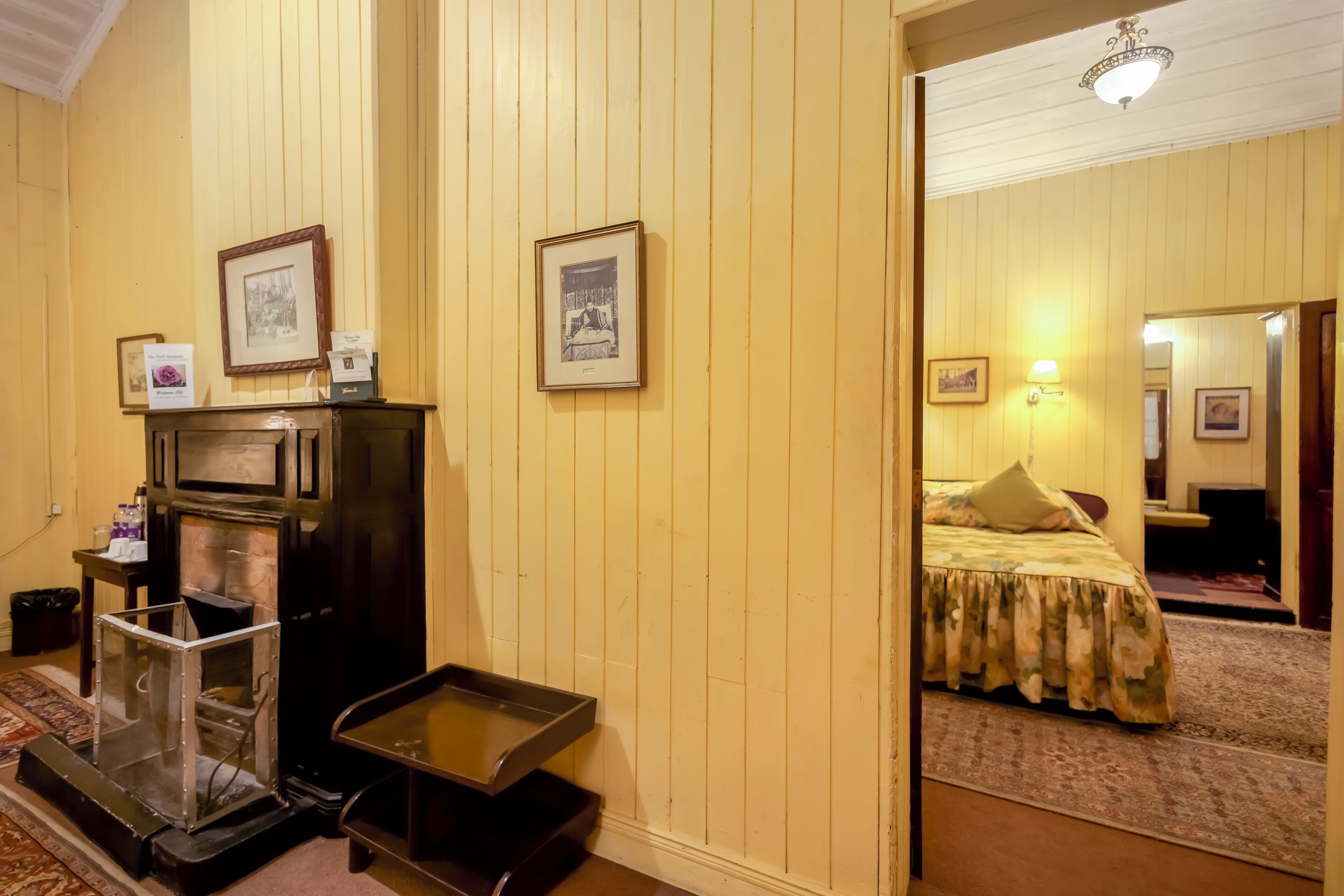 Une chambre d’hôtel avec un mur lambrissé en bois, un lit recouvert d’une courtepointe à motifs floraux, une petite table avec un plateau, et un tableau encadré accroché au mur.