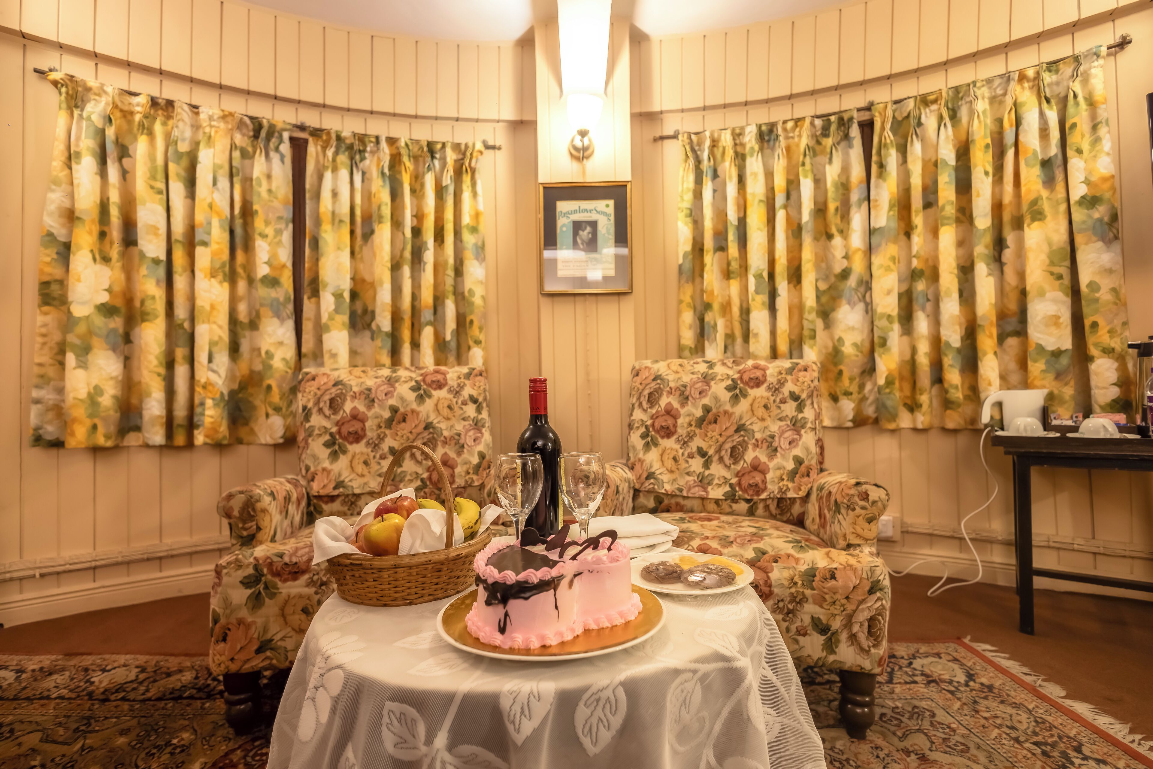 Una habitación con cortinas florales, dos sillones tapizados, una mesa con un pastel, una botella y una cesta de fruta.