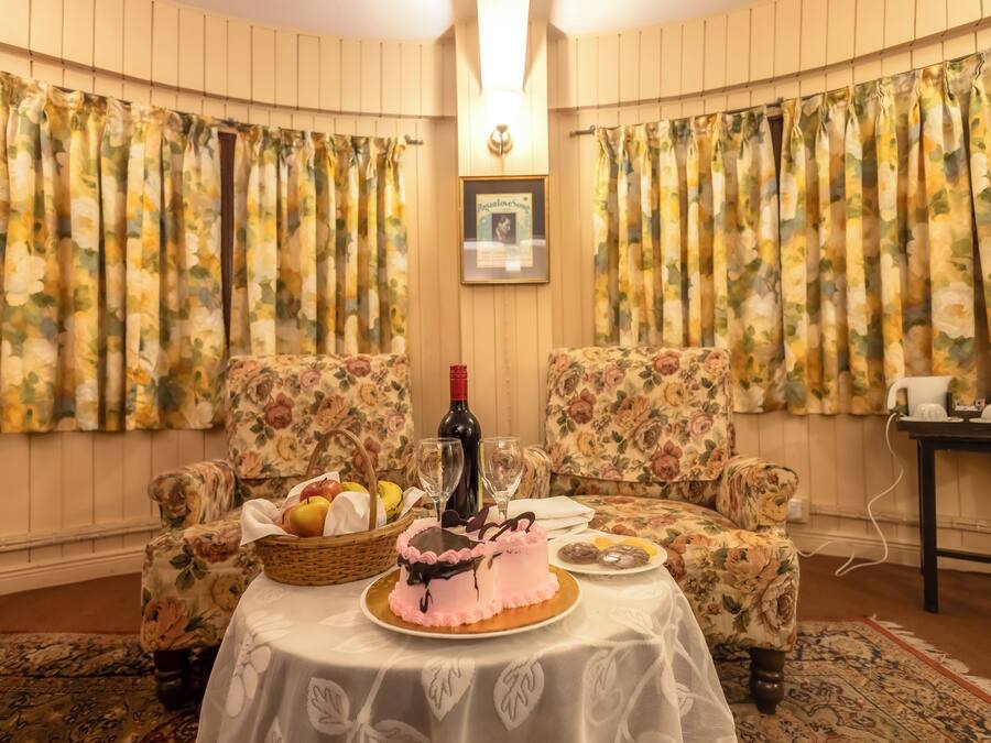 Una habitación con cortinas florales, dos sillones tapizados, una mesa con un pastel, una botella y una cesta de fruta.