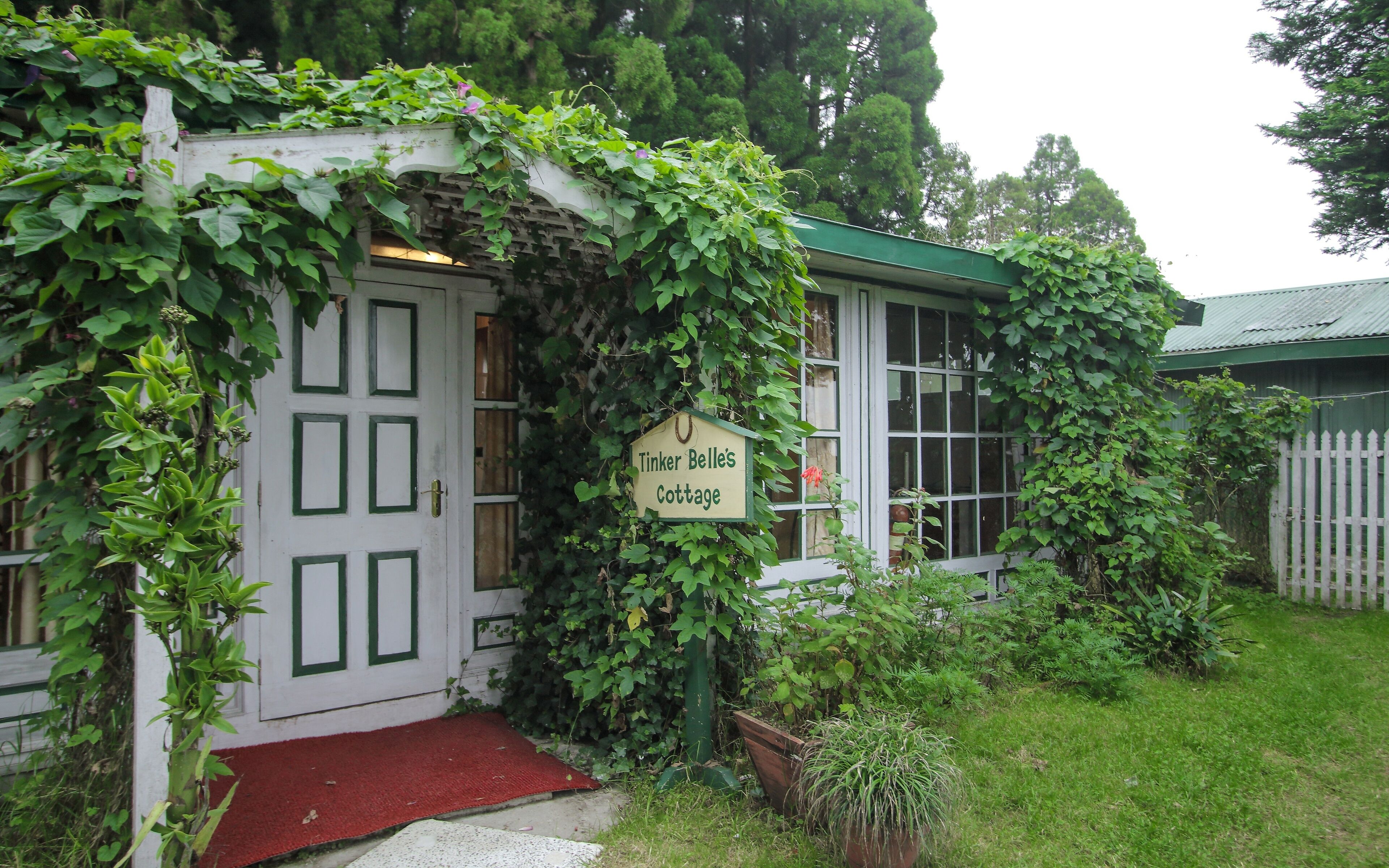 A cottage with a sign reading "Tinker Belle's Cottage," surrounded by lush greenery and a red mat at the entrance.