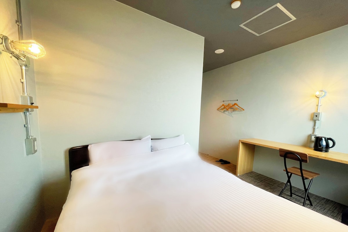 A hotel room with a large bed, a desk, and a chair. There is a wall-mounted light fixture above the bed and a hanging light above the desk.