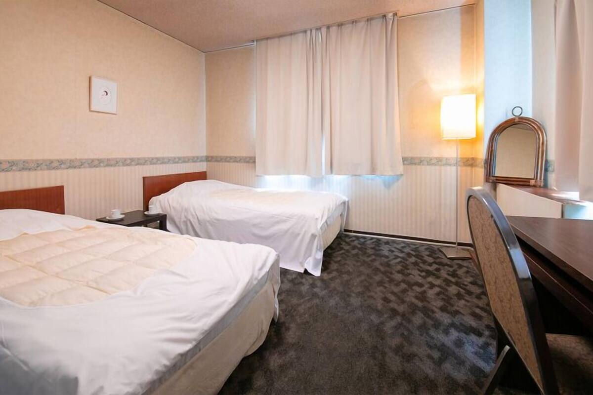 A hotel room with two beds, a desk, a chair, a lamp, and a window with curtains.