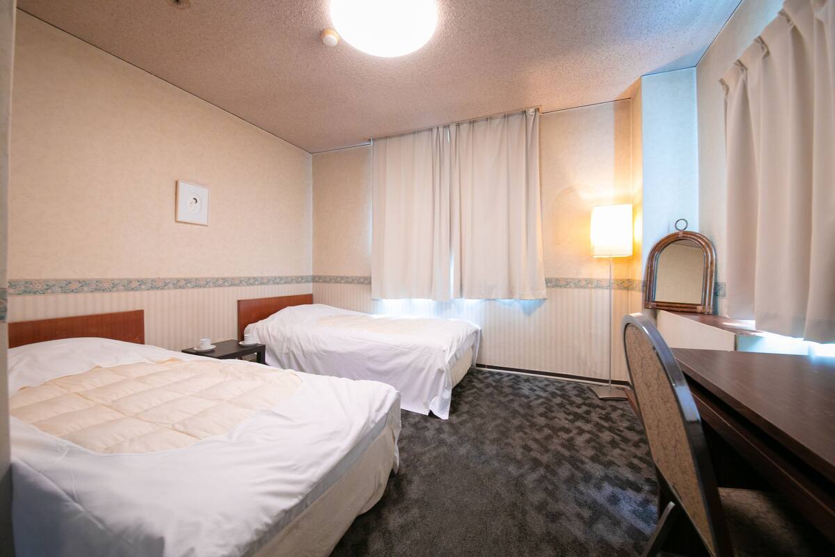 A hotel room with two single beds, a desk with a chair, a mirror, a lamp, and a window with curtains.