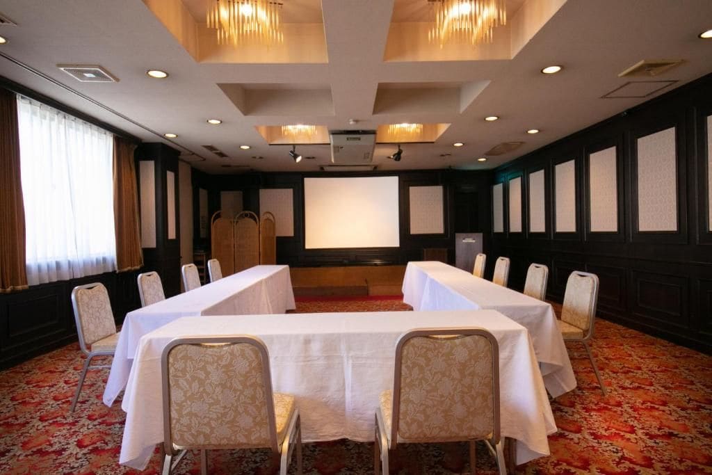 Un salón de conferencias con una larga mesa, sillas, una pantalla de proyector y piso alfombrado.