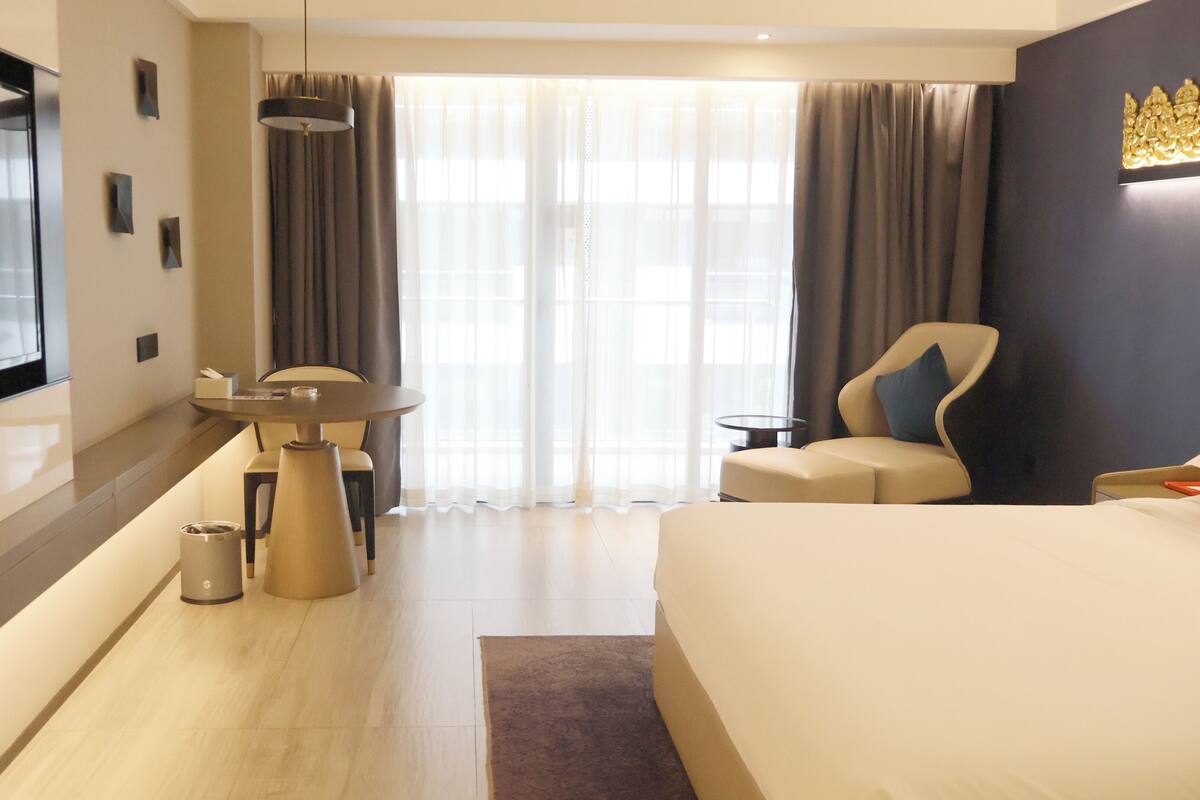 Ein Hotelzimmer mit Bett, Schreibtisch, Sessel, Fernseher und einem großen Fenster mit Vorhängen.