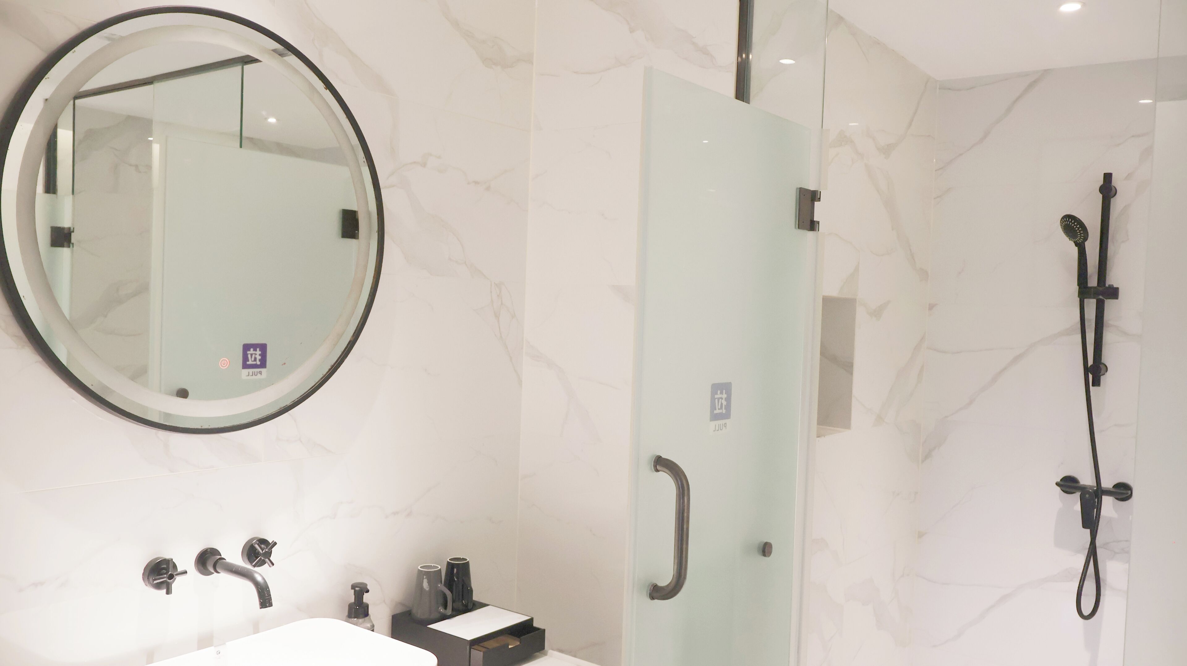 Une salle de bain moderne avec un grand miroir rond, une douche à parois de verre, une douchette à main et un lavabo avec un robinet.