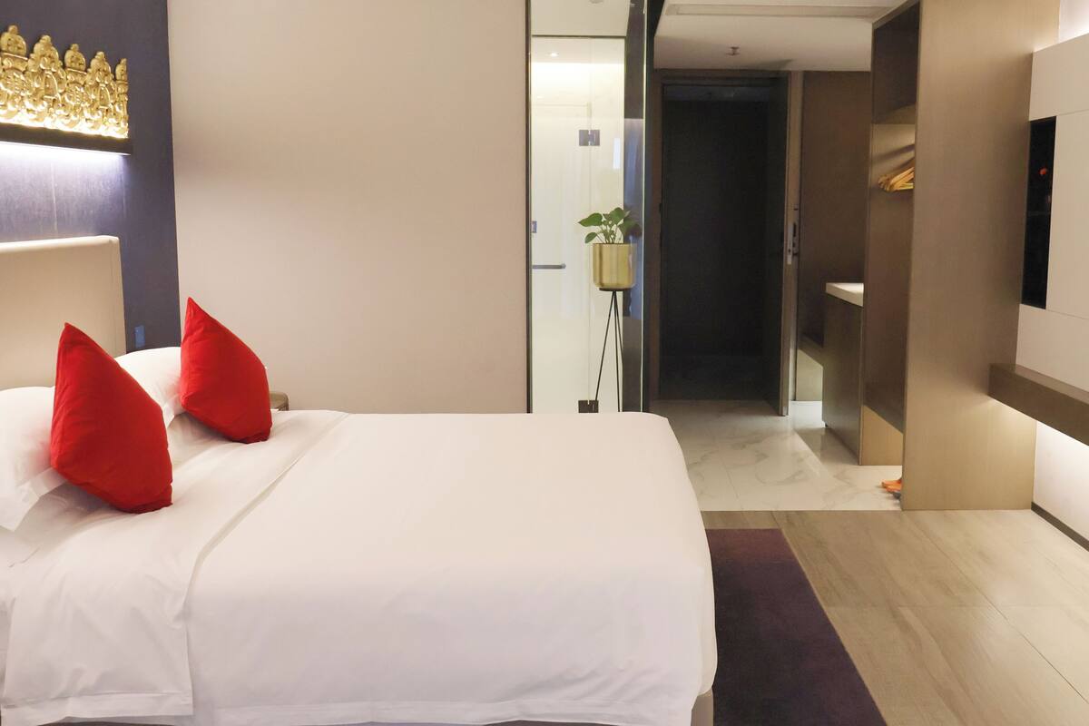 Ein Hotelzimmer mit Bett, roten Kissen, Fernseher, Pflanze und einem zur nächsten Tür führenden Flur.