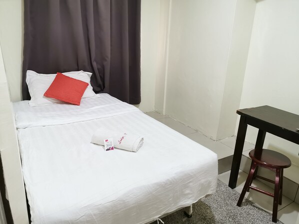 A neatly made bed with white linens, a red pillow, and a folded towel with a tag. A small wooden stool is beside the bed.