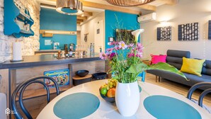 A modern kitchen with a blue backsplash, a dining area with a white table and blue placemats, and a living room with a gray sofa and colorful cushions. - Casa Amando Eco Boutique House (Rovinj)